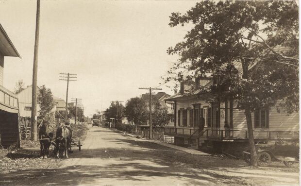 Photo en noir et blanc d'une rue en terre battue, avec des maisons de chaque côté.. Sur la gauche, deux chevaux attelés à une charette et sur la droite, une voiture d'antan. 