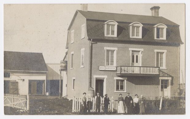 Photo en noir et blanc de neuf personnes devant une maison de briques de trois étages. Au-dessus de la porte est inscrit : « Hôtel Marcotte ».