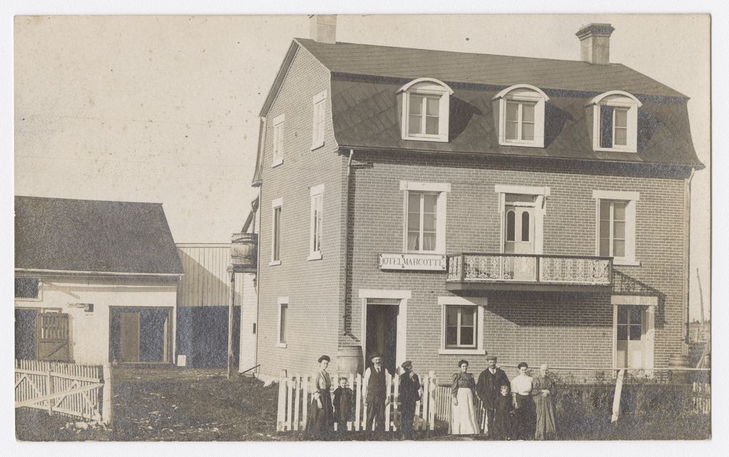 Photo en noir et blanc de neuf personnes devant une maison de briques de trois étages. Au-dessus de la porte est inscrit : « Hôtel Marcotte ».