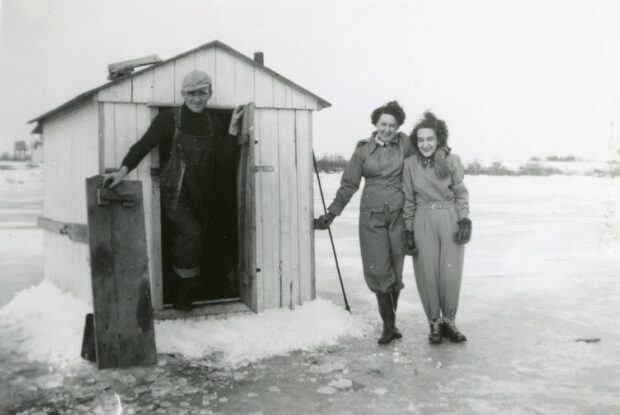 Photo en noir et blanc de deux femmes et un homme à côté d'une cabane de pêche sur glace.