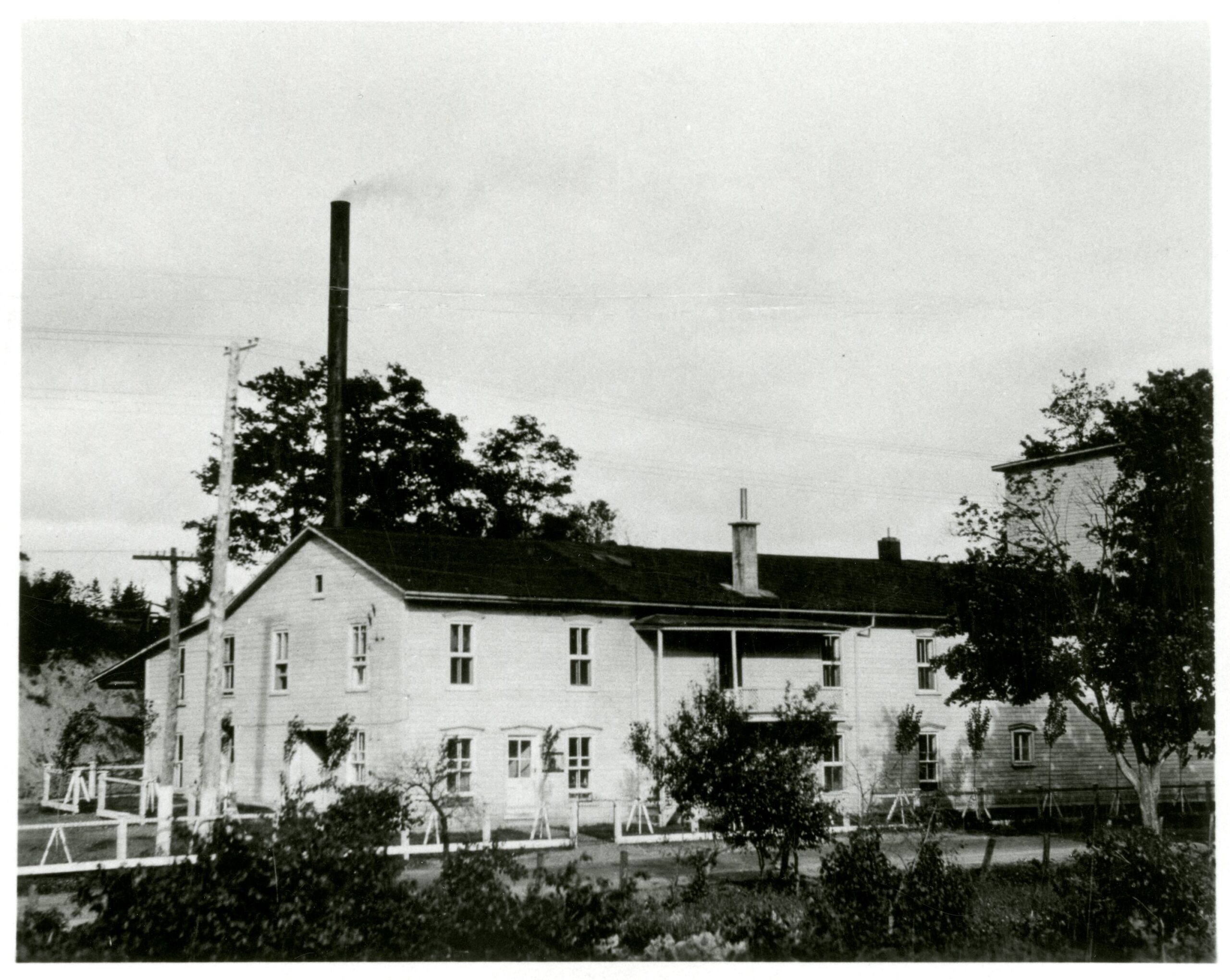 Photo en noir et blanc d'un long bâtiment de deux étages avec une cheminée haute. 