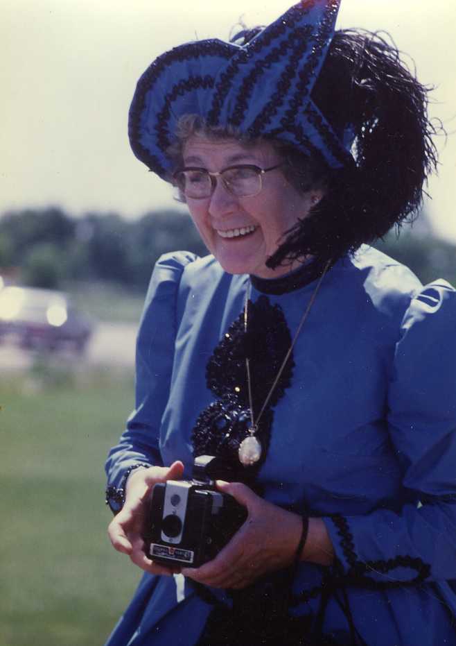 Thérèse Sauvageau vêtue d'une robe historique bleue tenant un appareil photo. 