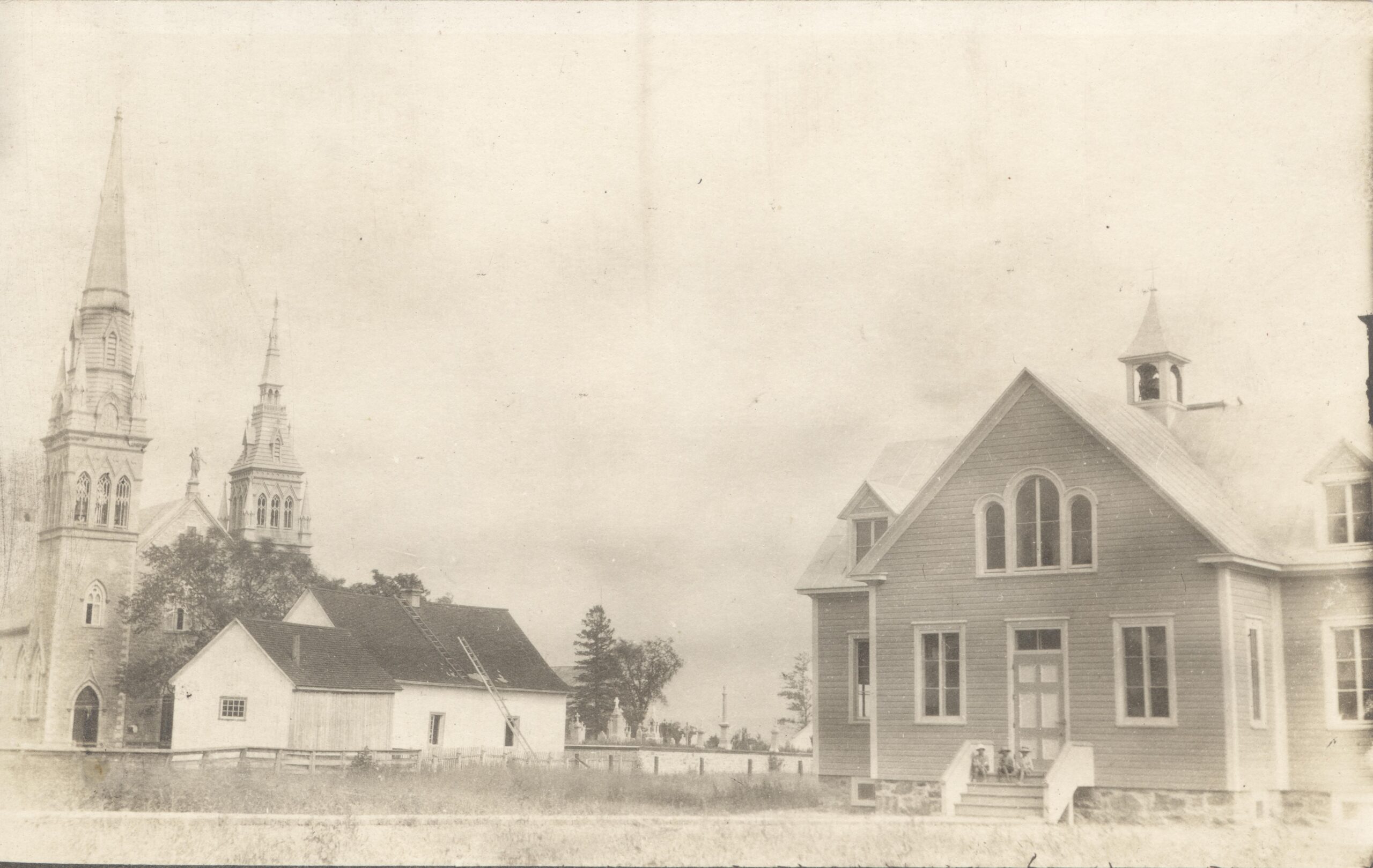 Photo en noir et blanc sur laquelle on aperçoit l'école (droite), la salle paroissiale (centre) et l'église (gauche). 