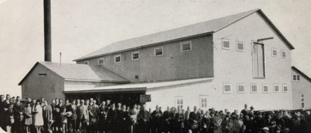 Photo en noir et blanc d'un groupe de personnes devant une usine de deux étages.