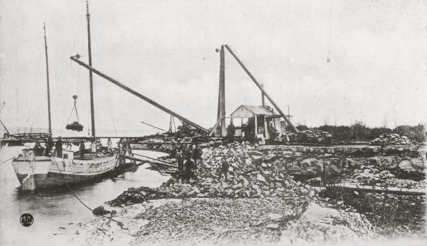 Photo en noir et blanc d'une carrière sur le bord de l'eau et d'un bateau en chargement. 