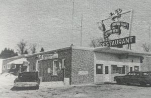 Black and white photo of a building with a badge reading “Restaurant” and two cars parked in front.