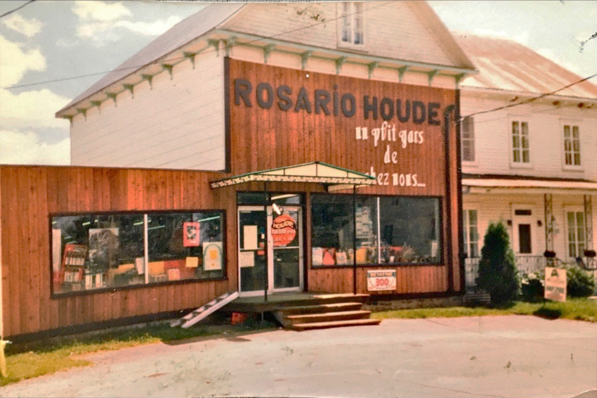 Photo d'une grande maison deux étages composée de deux bâtiments. Le bâtiment de gauche est occupé par un magasin général. Sur la devanture, il est inscrit "Rosario Houde - un p'tit gars de chez nous...". 