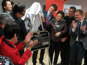 Un groupe de personnes dans une salle, dont des membres de l'Association Wong et Justin Trudeau, applaudissent lors du dévoilement d'une plaque commémorative.