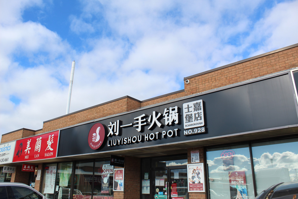 Un bâtiment d'un étage en briques rouges avec une enseigne noire, un logo rouge et des caractères chinois blancs et des lettres anglaises indiquant Liuyishou Hot Pot au-dessus de fenêtres en verre sur lesquelles sont apposées des affiches promotionnelles.