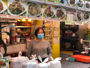 Une femme souriante portant un masque se tient derrière le comptoir d'un stand de nourriture très fréquenté, entourée de photographies de plats et de sacs à emporter, avec un panneau indiquant le certificat de l'inspection sanitaire.