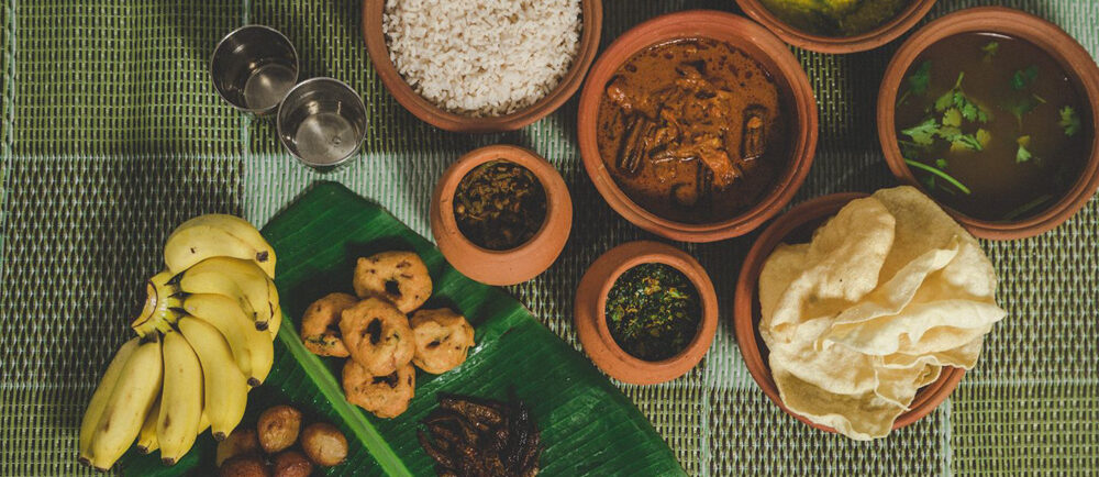 Une table sur laquelle sont étalés divers plats traditionnels, avec du riz, des currys et des bananes sur une natte tissée verte. Les plats sont servis dans des pots en terre cuite.