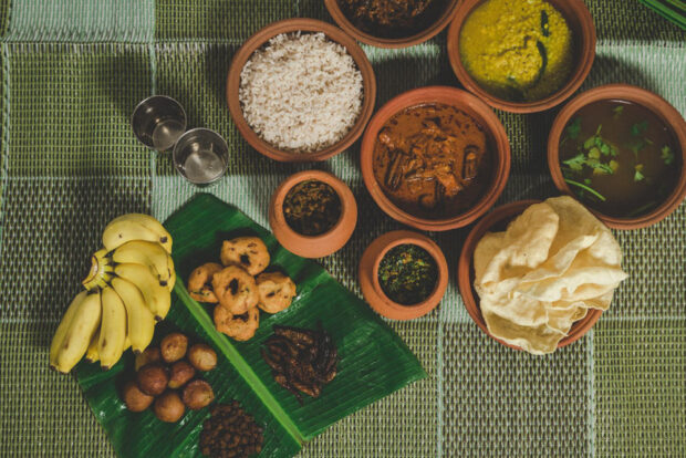 Une table sur laquelle sont étalés divers plats traditionnels, avec du riz, des currys et des bananes sur une natte tissée verte. Les plats sont servis dans des pots en terre cuite.