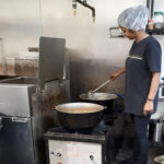 Une femme dans une cuisine, remuant une grande casserole sur une cuisinière industrielle. Elle porte un filet à cheveux et des vêtements décontractés, dans une cuisine aux surfaces métalliques.