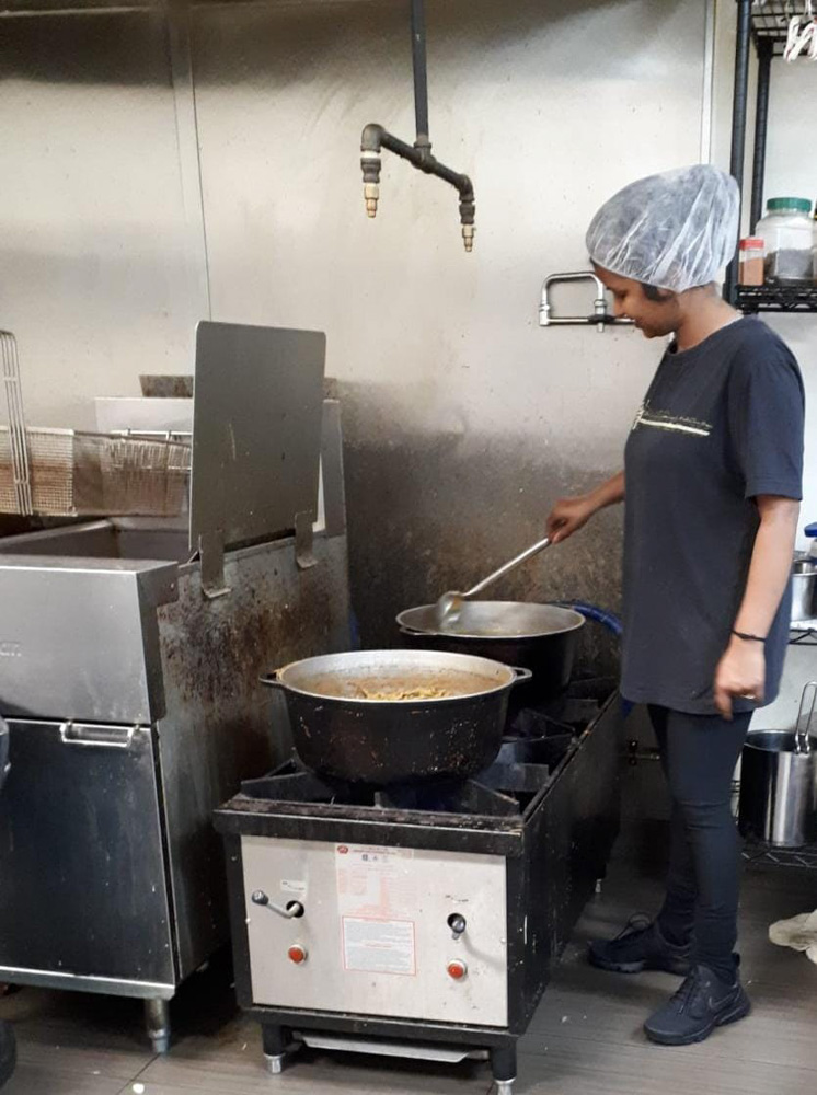 Une femme dans une cuisine, remuant une grande casserole sur une cuisinière industrielle. Elle porte un filet à cheveux et des vêtements décontractés, dans une cuisine aux surfaces métalliques.