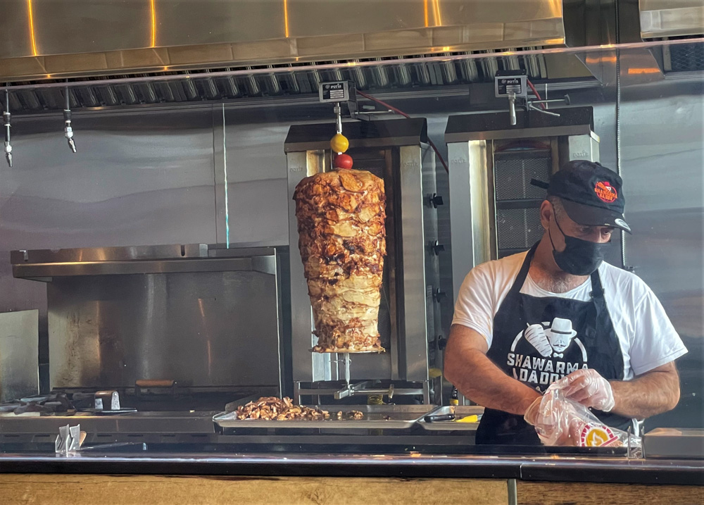 Un homme portant un masque et une casquette, ainsi qu'un tablier sur lequel on peut lire « Shawarma Daddy », découpe la viande au grand stand de shawarma rôti dans la cuisine d'un restaurant, entouré de surfaces métalliques et d'équipements de cuisson.