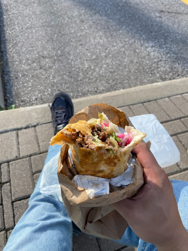 Une personne tenant un sandwich au chawarma roulé avec de la viande, des légumes et de la sauce visibles. Ils sont à l'extérieur, avec la surface pavée du stationnement en dessous d'eux.