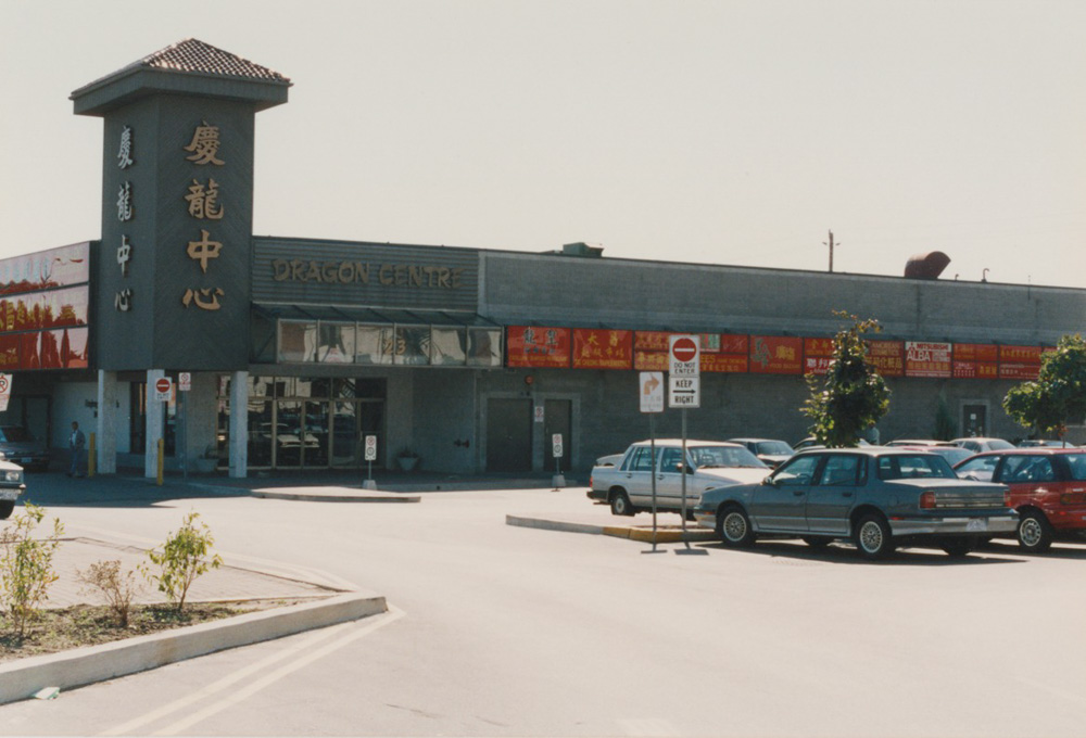 Bâtiment d'un étage en briques grises avec des enseignes de magasins, une tour en briques grises avec des caractères chinois stylisés et des voitures garées dans un grand stationnement.