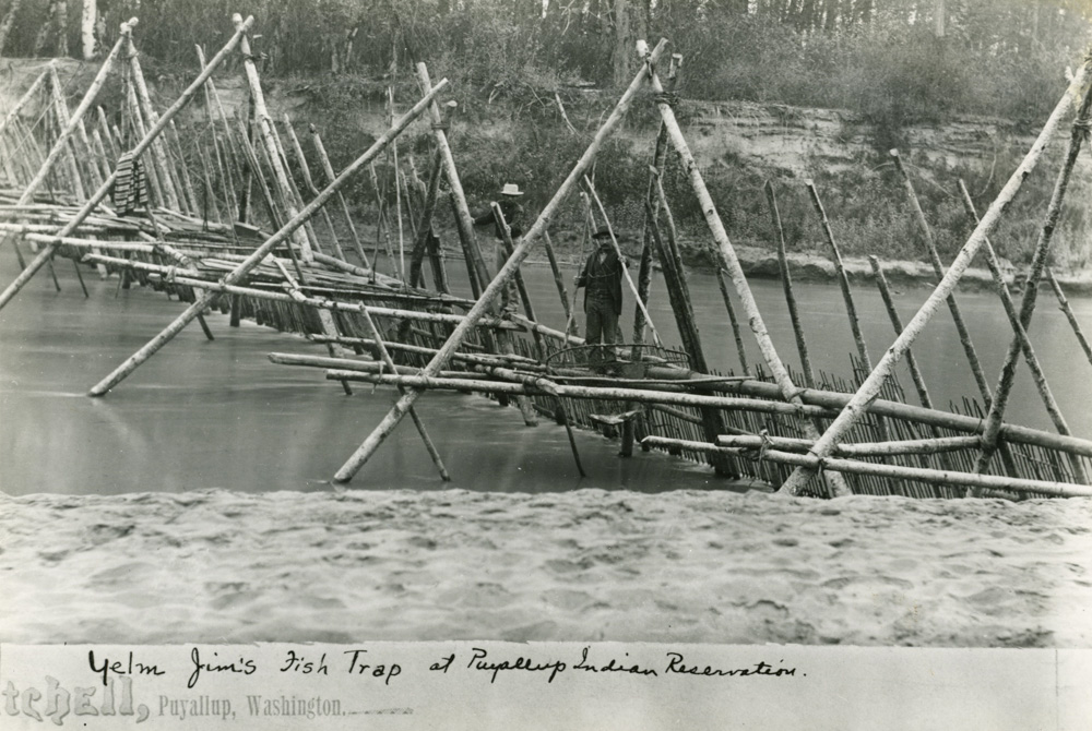 Une photographie en noir et blanc d'une structure de barrage de pêche, faite de longs bâtons de bois disposés en triangle sur un pont de bois qui s'étend sur une rivière, avec deux hommes debout sur la structure du pont.