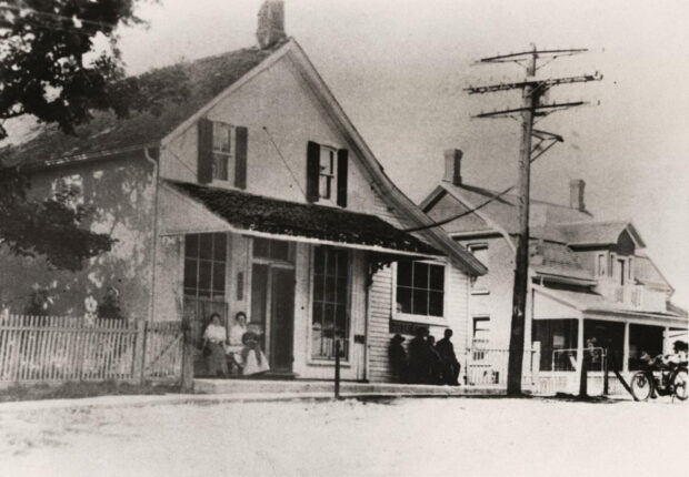 Une photographie en noir et blanc représentant une maison de ville blanche de deux étages avec deux petites fenêtres au deuxième étage, deux grandes fenêtres au rez-de-chaussée sous une véranda, séparées par une grande porte dans une large rue avec d'autres maisons, une clôture de piquets, des arbres et une grande ligne à haute tension. Devant les fenêtres sont assis une femme, deux jeunes enfants à gauche, et quatre silhouettes sombres à droite.