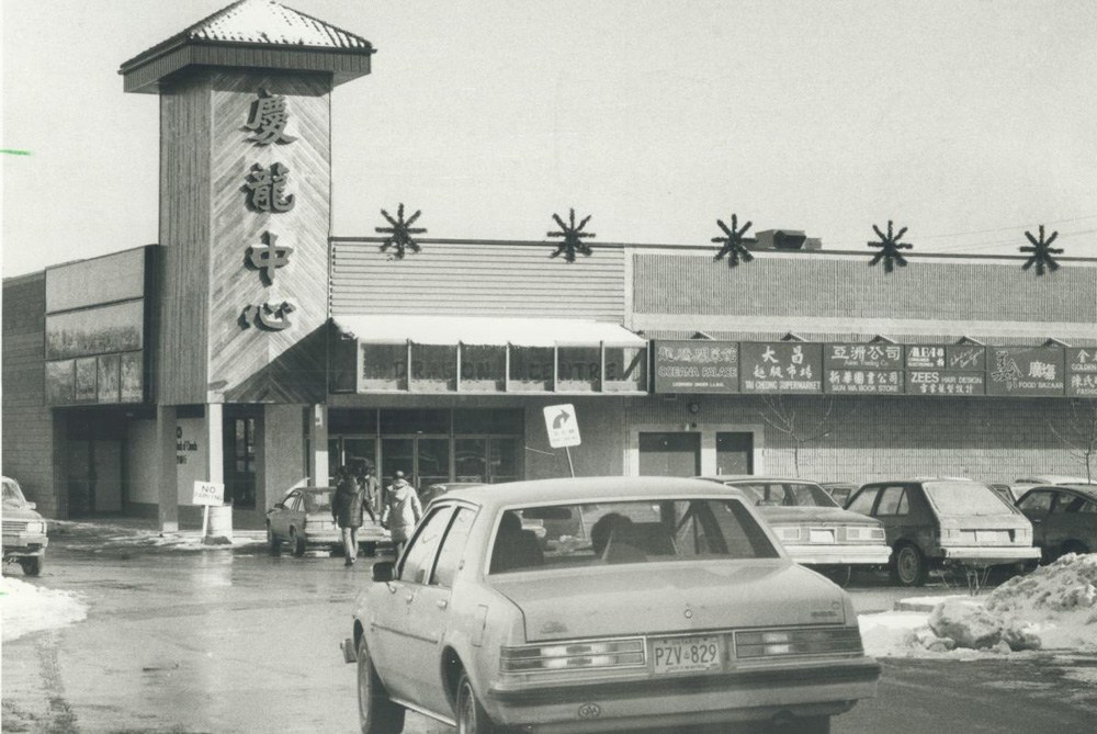 Une image en noir et blanc du Dragon Centre, appelé à l'époque le New Chinese Mall, avec des enseignes de magasins, une tour avec des caractères chinois stylisés et un stationnement rempli.