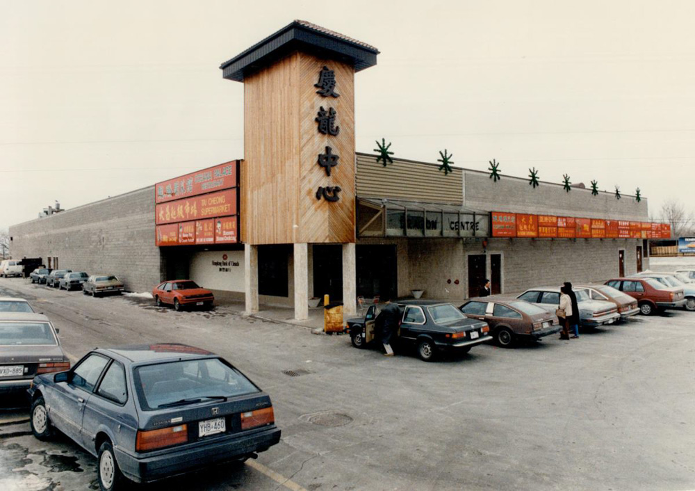 Une photographie analogique montrant le Dragon Centre Mall avec une façade en briques grises, une tour en bois avec des caractères chinois stylisés noirs, des enseignes de magasins rouges, et un stationnement plein avec des clients entrant dans le centre commercial.