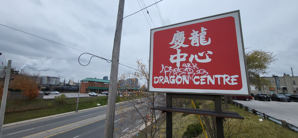 Une photographie en couleur d'un grand panneau rouge situé au sommet d'une route à six voies, sur lequel on peut lire Dragon Centre en anglais et en caractères chinois.