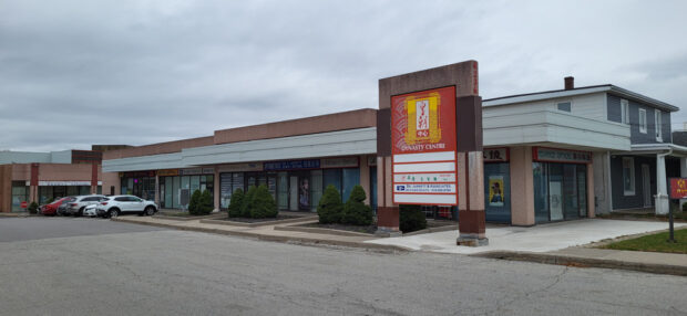 Une photographie en couleur de l'une des entrées pour voitures du Dynasty Centre Mall à Scarbrough montrant une large route, des voitures garées devant une rangée de commerces d'un étage avec une grande enseigne en rouge avec un texte lisant Dynasty Centre en blanc avec un logo stylisé jaune.