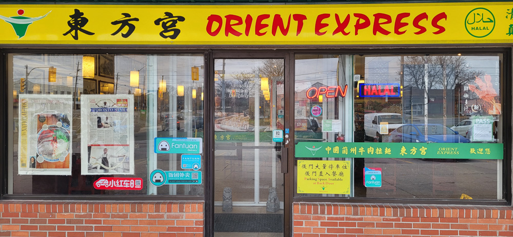 Une photographie en couleur de la façade du restaurant Orient Express à Torchin Plaza, Scarborough, montrant l'enseigne du restaurant en jaune avec un texte en rouge, une fenêtre montrant l'intérieur du restaurant et d'autres enseignes dans la fenêtre.