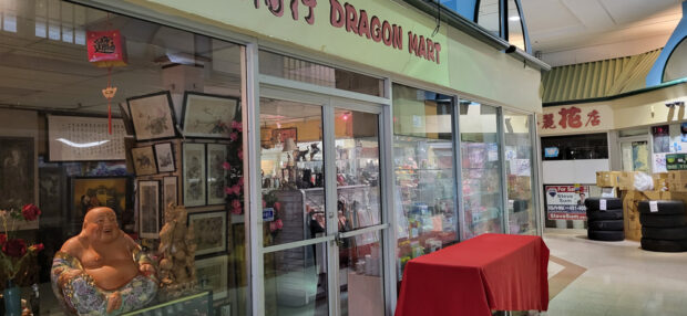 Devanture du « JOYS DRAGON MART » dans un centre commercial, présentant des statues traditionnelles chinoises, de l'art et des décorations rouges, avec un panneau « A vendre ».