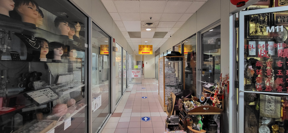 Un corridor intérieur d'un centre commercial avec des vitrines en verre présentant des perruques, des textiles et des objets culturels, menant à une aire de restauration.