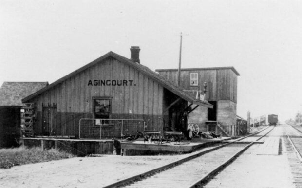 Une photo en noir et blanc d'une structure ressemblant à une grange avec un texte noir de grande taille indiquant Agincourt. La structure se trouve à côté de voies ferrées et représente l'une des premières gares d'Agincourt.