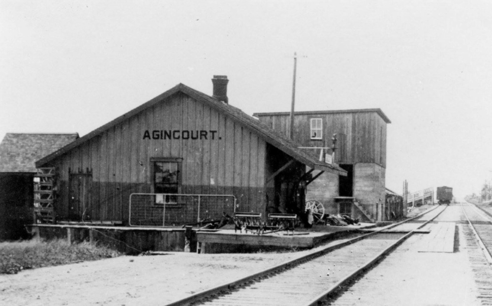 Une photo en noir et blanc d'une structure ressemblant à une grange avec un texte noir de grande taille indiquant Agincourt. La structure se trouve à côté de voies ferrées et représente l'une des premières gares d'Agincourt.