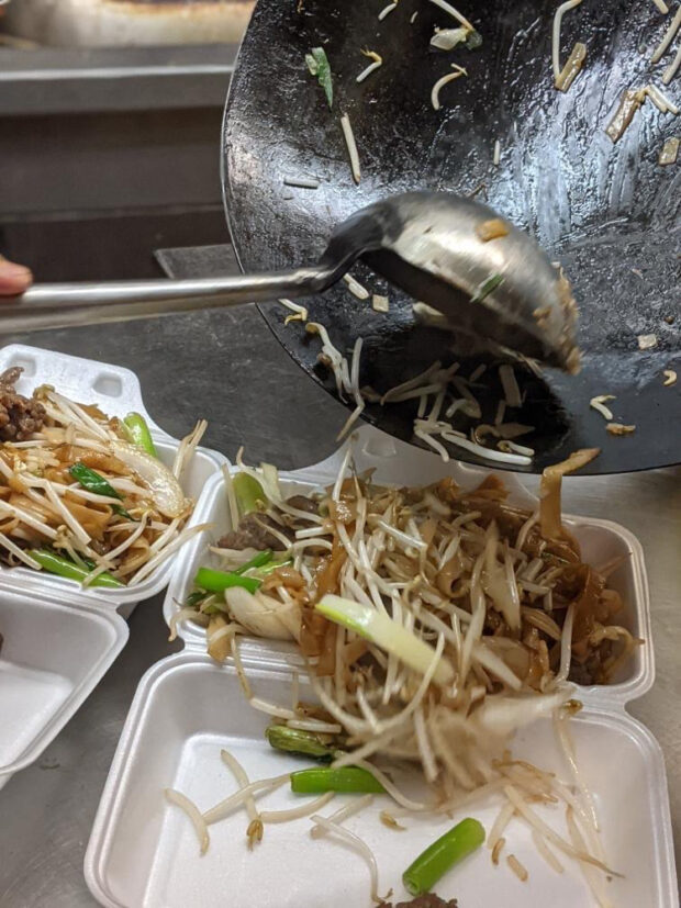 Un plan d'action d'un chef cuisinier transférant un sauté de germes de soja, d'oignons verts et de lanières de bœuf ou de porc d'un wok à des conteneurs en styromousse blanche.