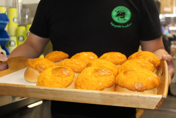 Un gros plan sur des brioches à l'ananas fraîchement cuites, qui sont des pains ronds avec une croûte jaune vif, présentées sur un plateau en bois, tenues par une personne portant une chemise noire avec un logo vert.