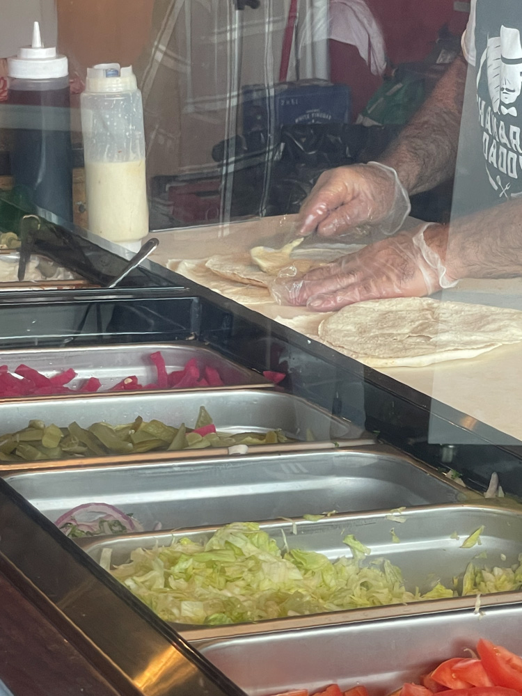 Une image en gros plan d'une personne préparant un sandwich au chawarma roulé derrière une cloison en verre, étalant une sauce de couleur crème sur un pain plat avec des plateaux de garnitures visibles au premier plan, y compris des cornichons, de la laitue et des tomates.