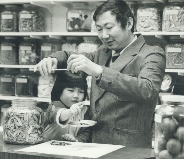 Une photographie en noir et blanc d'un Chinois en costume tenant une balance manuelle en équilibre sur un plan de travail. À côté de lui, un jeune enfant touche la balance. À gauche, un grand bocal en verre contenant des matières organiques et, dans un magasin, de nombreuses étagères de bocaux similaires contenant d'autres matières organiques.