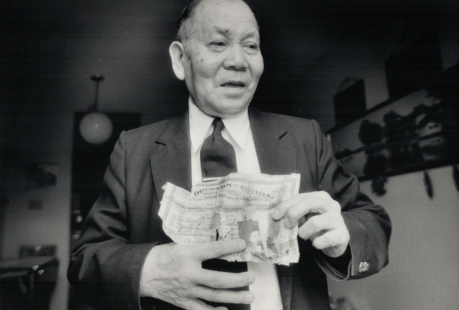 Un homme chinois d'un certain âge, en costume et cravate noirs, tenant un certificat vieilli avec des larmes et des inscriptions, photographié dans une maison.