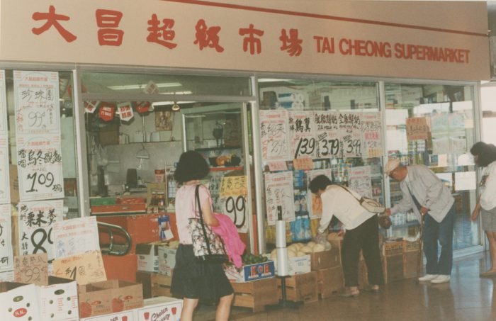 Une photographie en couleur d'un supermarché situé à l'intérieur d'un centre commercial, avec de grandes vitrines sur lesquelles sont apposées de nombreuses affiches manuscrites indiquant les prix des produits, des boîtes en carton contenant des produits alimentaires disposées sur le sol devant les vitrines et des clients locaux choisissant des produits dans les boîtes.