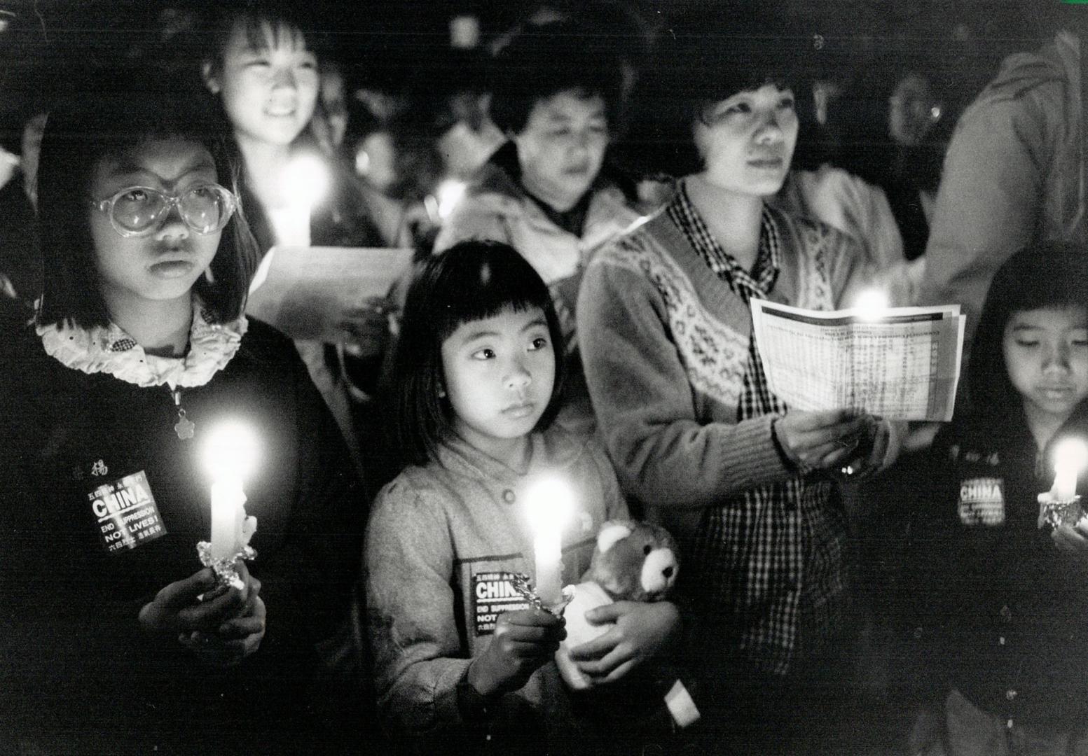 Une photographie en noir et blanc d'un rassemblement nocturne de femmes et de jeunes filles chinoises dans un groupe tenant des bougies, des brochures et des animaux en peluche, avec un air sombre.