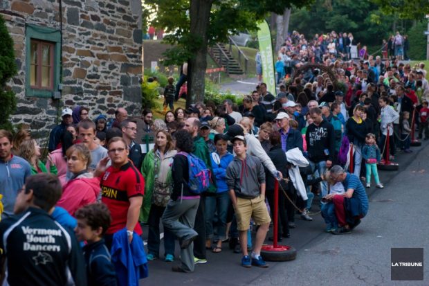 Photo couleur d’une longue file de personnes devant un bâtiment de brique ancienne qui se trouve à l’entrée du Parc de la gorge de Coaticook pour parcourir Foresta Lumina. 