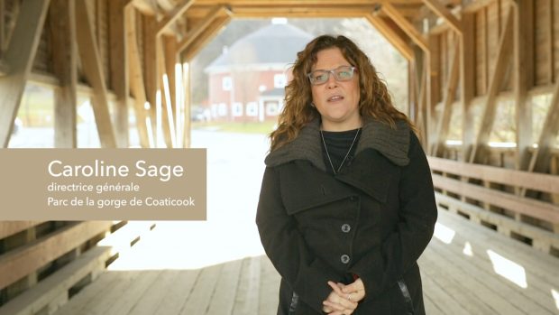 Photo couleur qui présente Caroline Sage, directrice générale de Parc de la gorge de Coaticook, habillée en manteau noir. Elle se tient sur le pont couvert du Parc de la gorge et derrière elle, on peut apercevoir la grange ronde, une bâtisse qui fait partie du parc.