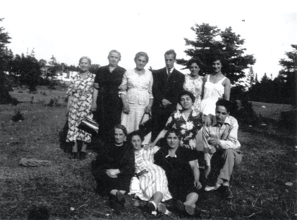 Photo d’extérieur en noir et blanc. Huit femmes et deux hommes sont rassemblés dans un champ avec des arbres en arrière-plan. Cinq femmes en robe se tiennent debout à l’arrière-plan avec un homme en costume entre elles; à l’avant, quatre femmes sont agenouillées ou assises sur le sol à côté d’un homme en chemise et pantalon.