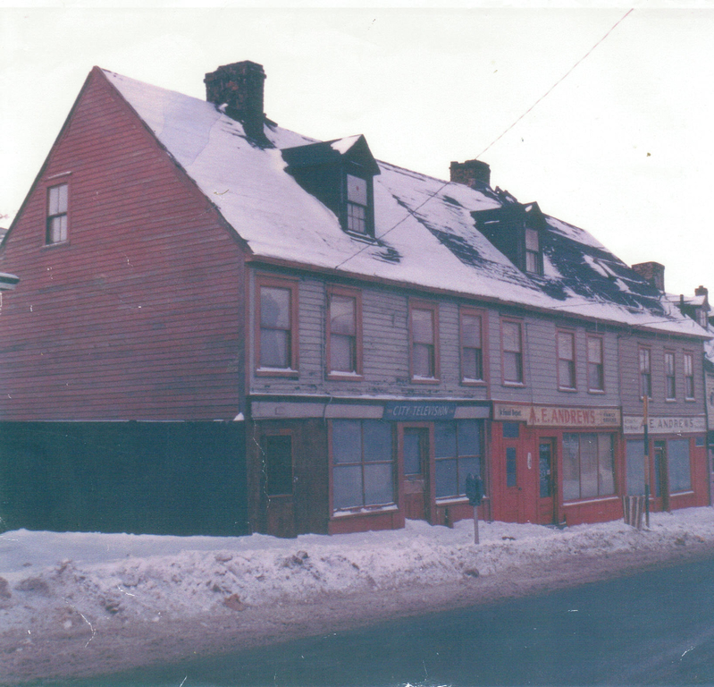 Paysage de rue en couleurs de trois maisons en rangée de trois étages. De gauche à droite, les enseignes au-dessus des entrées principales et des vitrines sont City Television, A. E. Andrews et AE Andrews. La maison de gauche est la plus grande, avec quatre fenêtres au deuxième étage, une lucarne au troisième étage et une cheminée. La maison du milieu a trois fenêtres au deuxième étage et une lucarne au troisième étage. La maison de droite a trois fenêtres au deuxième étage. Les maisons sont de couleur rouge et gris. Il y a une autre cheminée entre la maison du milieu et celle de droite.