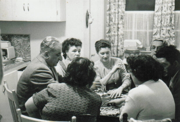 Photo d’intérieur en noir et blanc. Cinq femmes et deux hommes sont assis autour d’une table de cuisine et jouent aux cartes. L’une des femmes au centre de la photo fume.