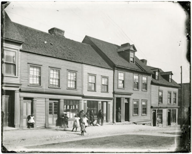 Photo d’extérieur en noir et blanc. Cinq maisons en rangée à l’arrière-plan. Six enfants jouent devant les maisons, quatre hommes sont adossés à une maison et une femme se tient dans l’embrasure de la porte d’une autre Maison.