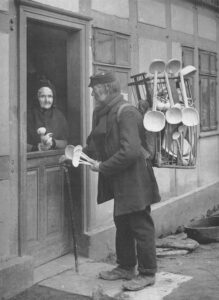 Photo d’extérieur en noir et blanc. Une femme est debout à sa porte et tient cinq cuillères. Un homme portant un chapeau, un manteau et un foulard tient une canne dans sa main droite et cinq cuillères en bois dans sa main gauche. Il se tient sur le pas de la porte de la femme avec un sac sur le dos rempli de cuillères et de louches en bois.