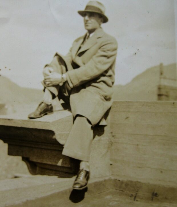 Portrait sépia. Un homme vêtu d’un chapeau mou, d’un pardessus, d’un pantalon et de chaussures habillées est assis sur le rebord d’un toit. Derrière l’homme, on peut voir la silhouette de deux collines.