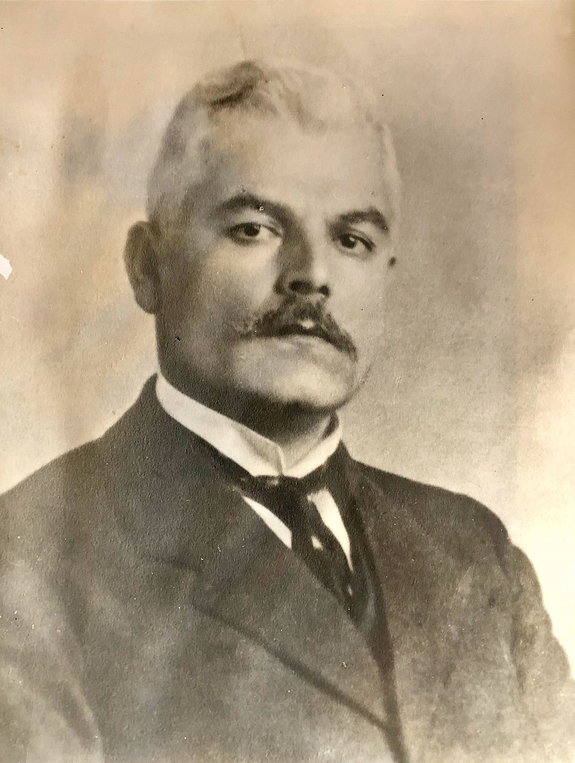 Portrait en noir et blanc d’un homme aux cheveux clairs, avec une moustache, des sourcils et des yeux foncés, portant un costume.