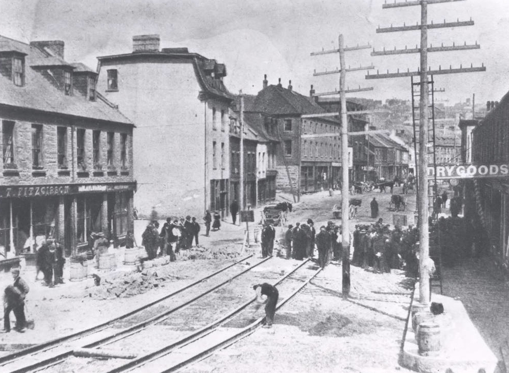 Paysage de rue en noir et blanc. Le côté gauche de la rue présente un mélange de maisons en rangée et de maisons attenantes devant lesquelles se trouvent de petits groupes de personnes. Des rails de tramway sont en cours d’installation au centre de l’image. Un gros groupe d’hommes se trouve sur la moitié droite de la photo, derrière deux poteaux électriques. À l’arrière-plan, il y a des charrettes tirées par des chevaux.