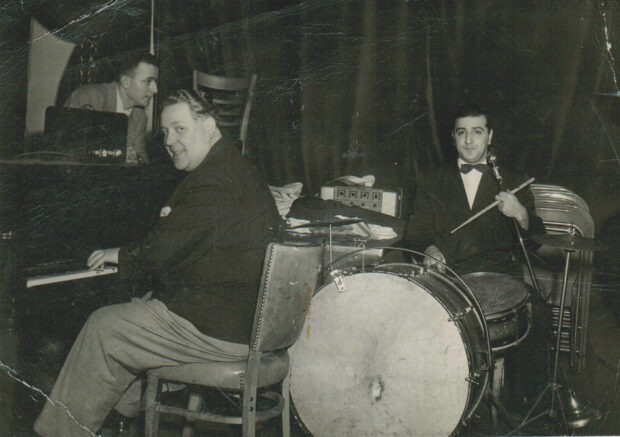 Photo d’intérieur en noir et blanc. Trois hommes avec des instruments de musique, dont un piano et une batterie. Les hommes et leurs instruments se trouvent devant un grand rideau.